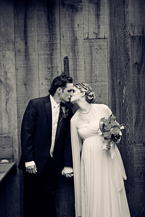 Black and white photo - Bride and groom holding hands and kissing in  front of rustic wall - Bride is wearing sheath style wedding dress with shawl and pearls -  Wedding photo by Meg Perotti
